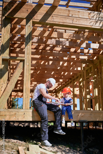 Father with toddler son constructing wooden frame house. Male builder and his son examining the construction plan, wearing helmets and overalls on sunny day. Carpentry and family concept.