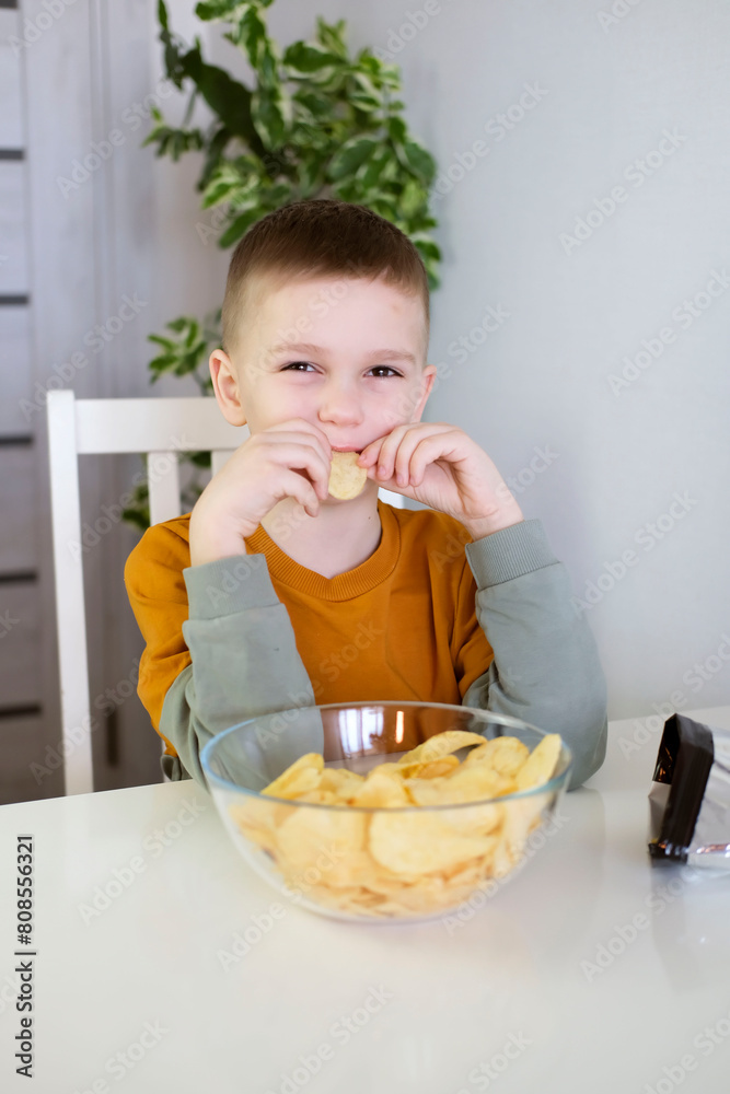 Hungry child happily eats potato chips while sitting at table. Boy eating fast food, junk food at home. Vertical photography. Hungry child happily eats potato chips while sitting at table. Boy eating fast food, junk food at home. Vertical photography.