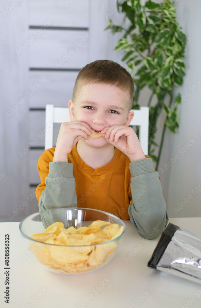 Hungry child happily eats potato chips while sitting at table. Boy eating fast food, junk food at home. Vertical photography. Hungry child happily eats potato chips while sitting at table. Boy eating fast food, junk food at home. Vertical photography.