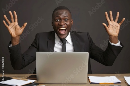 A man in a suit is sitting at a desk with a laptop open in front of him. He is smiling and he is excited