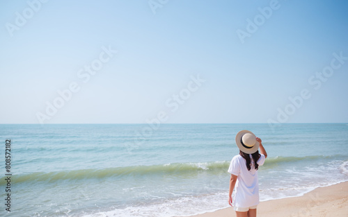 Rear view image of a young woman with hat standing on the beach with blue sky background