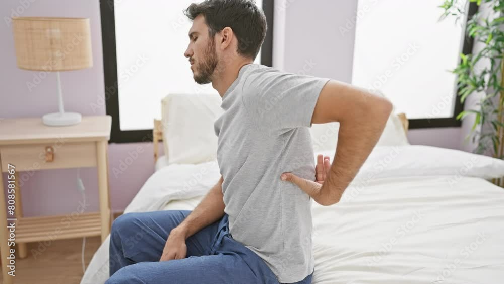 Young man experiencing back pain while sitting on bed in a well-lit bedroom, portraying a health issue in a domestic setting. Young man experiencing back pain while sitting on bed in a well-lit bedroom, portraying a health issue in a domestic setting.