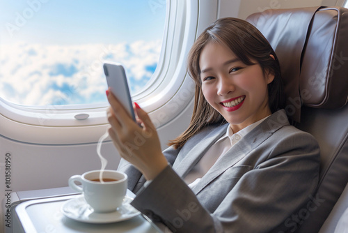 A happy Asian businesswoman in a business suit leans back in her airplane seat and takes a selfie with her phone.  photo