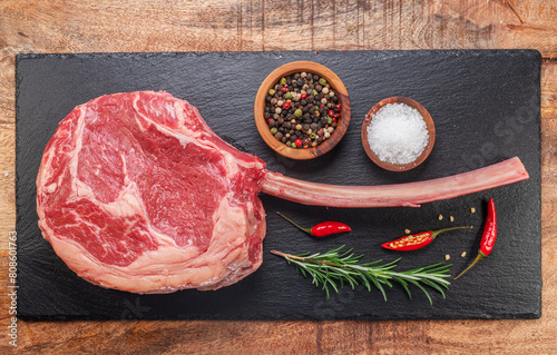 Raw rib steak with bone or tomahawk steak with seasonings on slate serving plate. Flat lay.