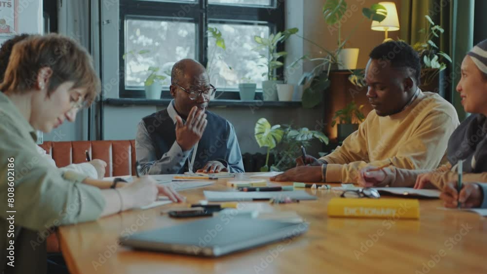 Mature Black teacher speaking to group of diverse immigrant students sitting around table and writing down notes during language class in school Mature Black teacher speaking to group of diverse immigrant students sitting around table and writing down notes during language class in school