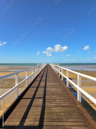 A long wooden pier stretching to the horizon, over a calm beach at low tide with clear blue sky, yellow sand and little white clouds.