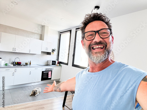 One man dow owner take selfie picture indoor inside house showing kitchen and background space. Happy mature male with beard and glasses smile at the camera and show home interior. Cheerful people