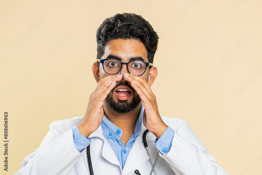 Keep my secrets, silence. Indian doctor cardiologist man whisper news rumors holding hands near mouth, share gossip, quiet. Arabian guy telling interesting confidential information on beige background Keep my secrets, silence. Indian doctor cardiologist man whisper news rumors holding hands near mouth, share gossip, quiet. Arabian guy telling interesting confidential information on beige background