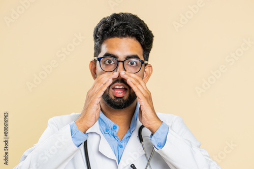Keep my secrets, silence. Indian doctor cardiologist man whisper news rumors holding hands near mouth, share gossip, quiet. Arabian guy telling interesting confidential information on beige background