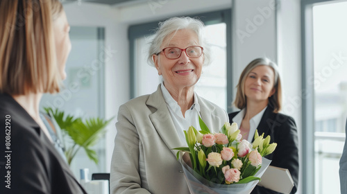 copy space  stockphoto  successful elderly business woman recieving a bouquet. multiracial team is standing around the businesswoman  modern office. Senior business woman going on retirement  pension.