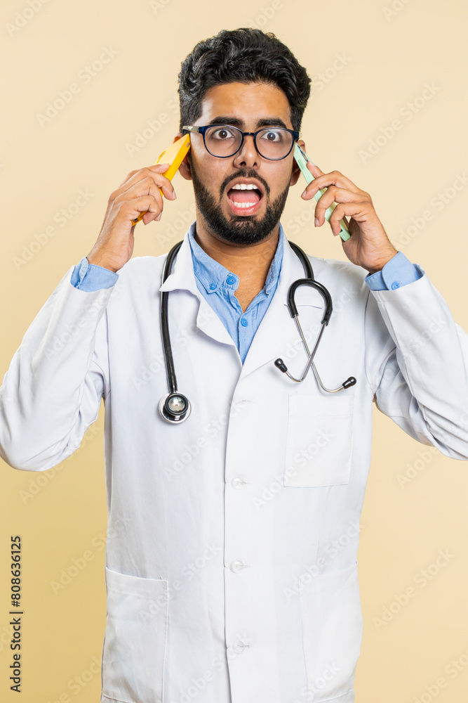 Irritated nervous stressed tired Indian doctor cardiologist man talking screaming on two mobile phones have conversation conflict quarrel. Arabian apothecary pharmacy guy on beige background. Vertical Irritated nervous stressed tired Indian doctor cardiologist man talking screaming on two mobile phones have conversation conflict quarrel. Arabian apothecary pharmacy guy on beige background. Vertical