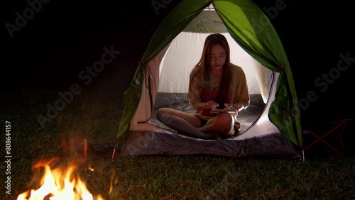 asiatic woman camping outdoor chatting inside her tent with modern smartphone connected at 5g in front of bonfire  photo