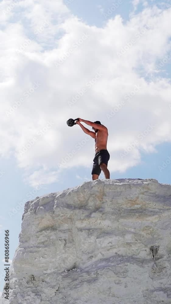Strong man lifting kettlebell on the mountain in summer. Male athlete swinging kettlebell with two hands on the beautiful natural background outdoors. Vertical video Strong man lifting kettlebell on the mountain in summer. Male athlete swinging kettlebell with two hands on the beautiful natural background outdoors. Vertical video