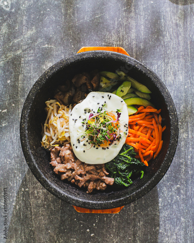 A vibrant Korean bibimbap served in a black stone bowl, featuring sections of minced meat, various julienned vegetables, a fried egg on top, sprinkled with sesame seeds photo