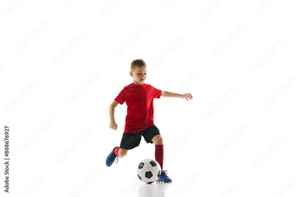Sporty little boy, kicks ball to make perfect shot against white studio background. Small football player makes perfect pass. Concept of professional sport, championship, youth league, hobby. Ad
