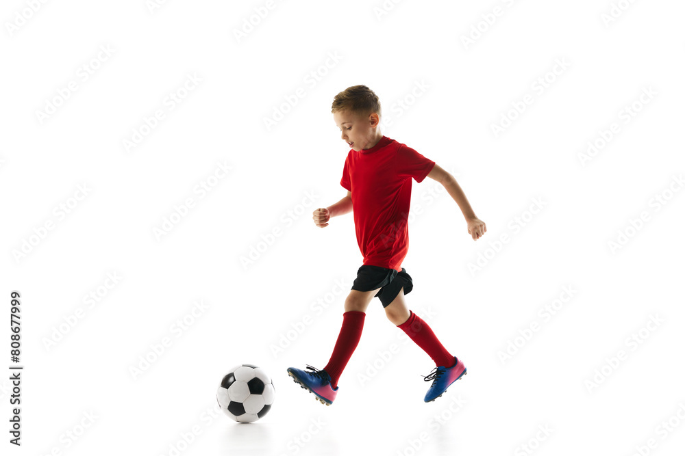 Little athlete boy, dribbling ball to take perfect goal against white studio background. Young soccer player in motion. Concept of professional sport, championship, youth league, hobby. Ad