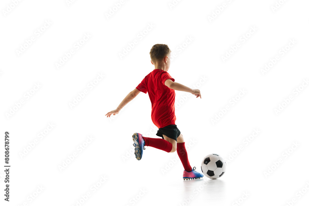 Sporty little boy, kicks ball to make perfect shot in motion against white studio background. Small football player makes perfect pass. Concept of professional sport, championship, youth league, hobby