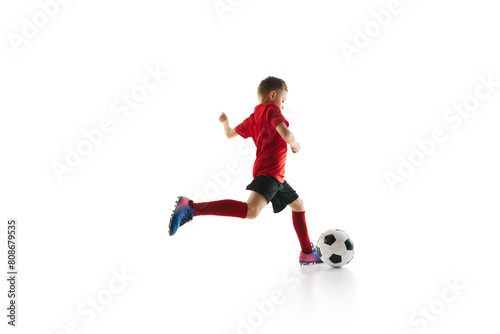 Sporty little boy, kicks ball to make perfect pass in motion against white studio background. Small football player makes goal. Concept of professional sport, championship, youth league, hobby. Ad