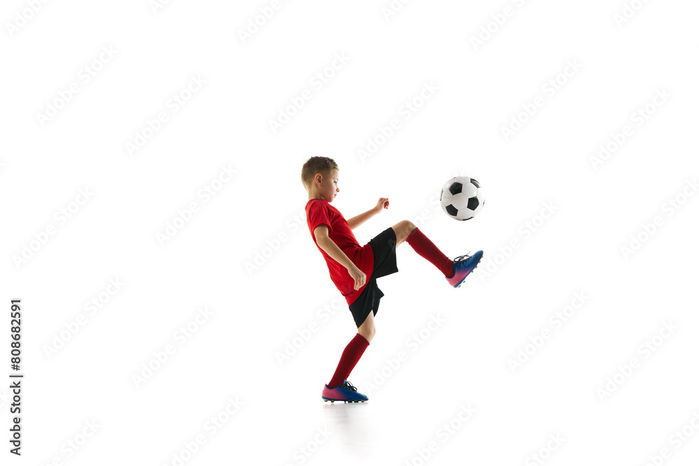 Dynamic portrait of little sporty boy with soccer ball doing flying kick in motion against white studio background. Concept of professional sport, championship, youth league, hobby. Ad