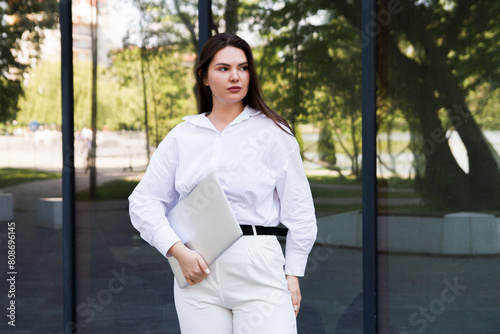 A woman in a park in a white shirt and white pants stands with a laptop in her hands near the office