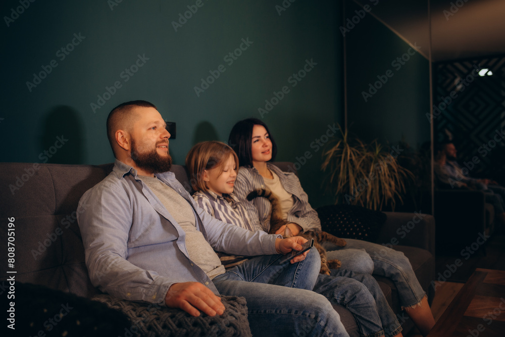 family, leisure and people concept - happy smiling father with remote control, mother and little daughter watching tv at home at night family, leisure and people concept - happy smiling father with remote control, mother and little daughter watching tv at home at night