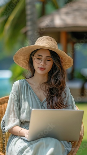 Dressed in a frock, sunglasses, and a hat, the Asian woman traveller is using her laptop to conduct business on the lawn in front of a stunning resort hotel.