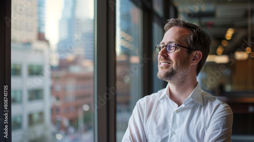 Portrait of Product manager happily looking contemplative out of an office window. He has a smile as he looks outside of the window at downtown office buildings. © Alina Tymofieieva