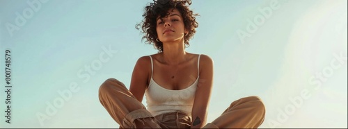 Woman with curly hair in a casual linen outfit sitting on the ground, clear facial features. She wears a white tank top and brown pants. A desert background with a white sky.