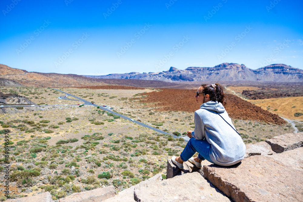 A girl sitting beneath Mount Teide, gazing out over the valleys of Gran Canaria, Tenerife. The majestic volcano looms in the background, while the vast landscape unfolds before her.