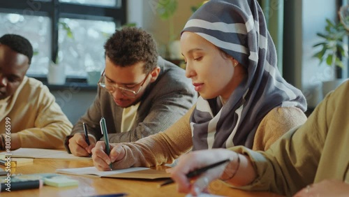 Young Muslim woman in hijab sitting with diverse group mates at desk in classroom and writing in copybook when studying in language school for immigrants photo