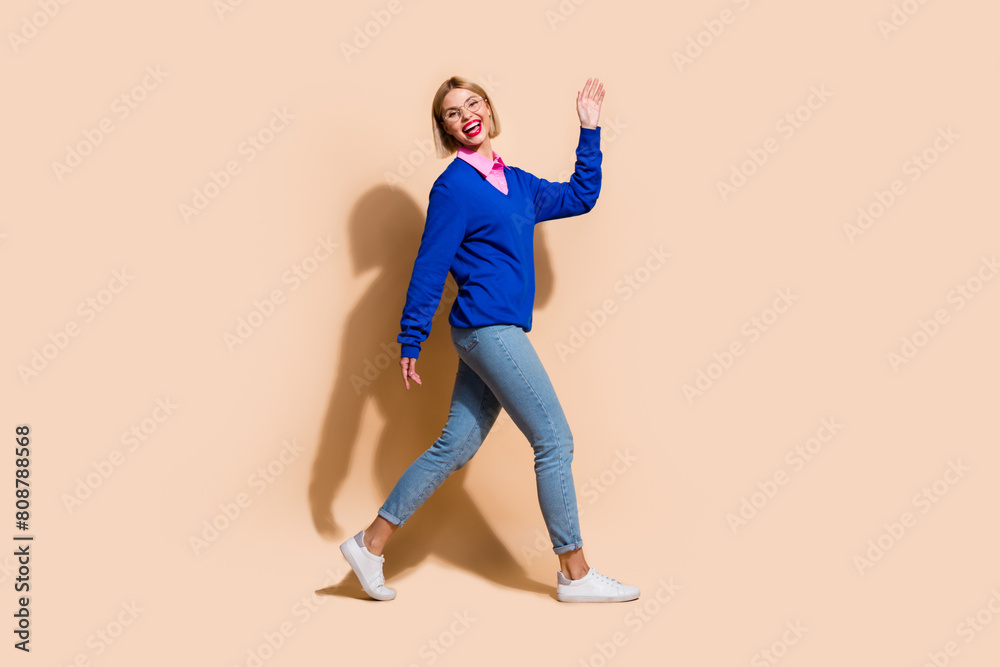 Full body profile photo of stunning cheerful lady arm waving hi walk empty space isolated on beige color background Full body profile photo of stunning cheerful lady arm waving hi walk empty space isolated on beige color background