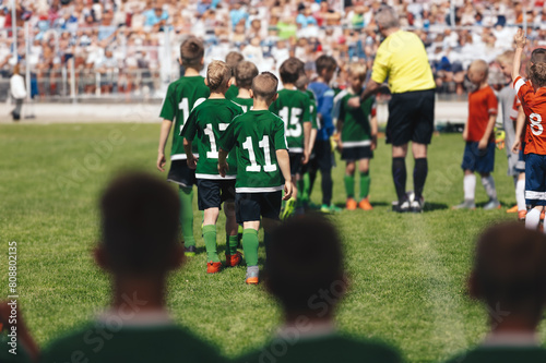 Children's football players walk onto the pitch with the referee. Kids play tournament matches. Schoolboys play the game on grass stadium. Spectators in the blurred background sitting on stadium seats