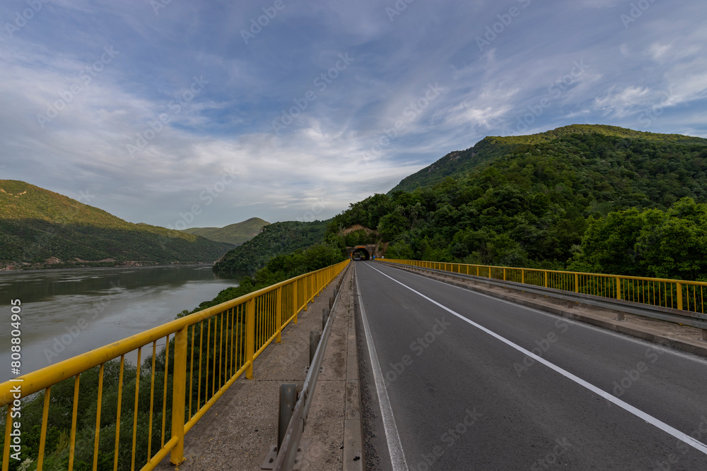 The river flows beneath, and the mountains in the background are lush with greenery, creating a peaceful and remote scene. The sunlight casts shadows on the road, The river flows beneath, and the mountains in the background are lush with greenery, creating a peaceful and remote scene. The sunlight casts shadows on the road,