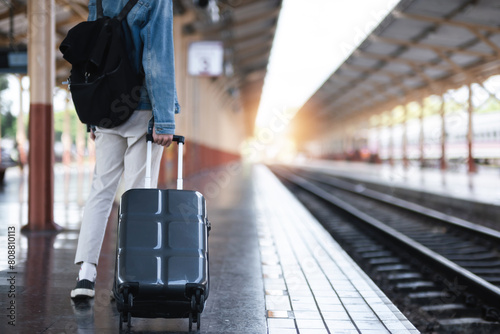 A young man holding a suitcase waits for a train at the train station for traveling.