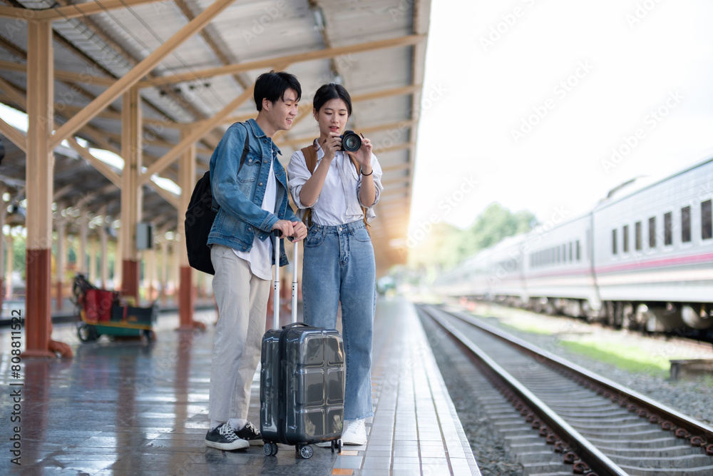 Happy young Asian couple carrying backpacks and cameras preparing to wait for the train at the train station waiting for their vacation trip together.