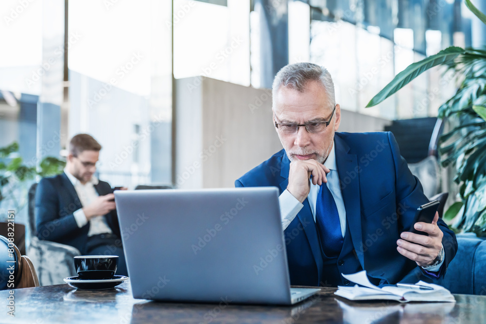Senior businessman investor entrepreneur working on laptop computer with mobile phone in hands at office lobby. Portrait of confident mature senior business man.