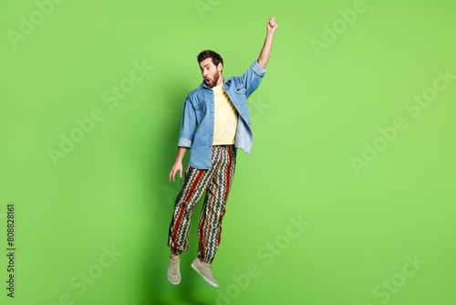 Full length photo of excited handsome guy wear denim jacket jumping high catching arm empty space isolated green color background photo