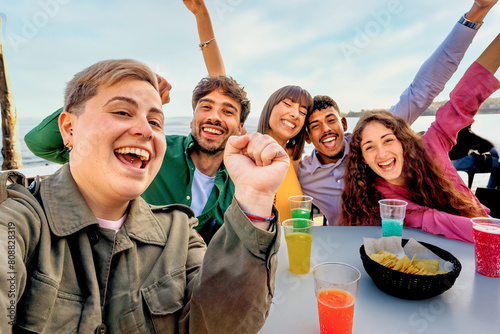 Group of diverse friends selfie - laughing and enjoying drinks at a beach gathering, showing joy and togetherness in a casual outdoor setting.