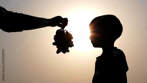 Silhouette views an Asian kid standing under the hot sun and eating grapes. Concept of eating fruits to prevent dehydration in the body.  photo