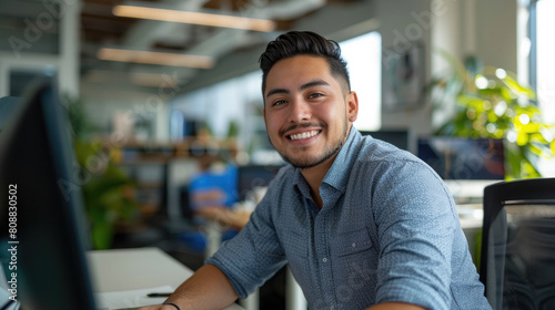 Happy and successful young latino male software engineer working at desk with computer looking front and smiling against modern office background