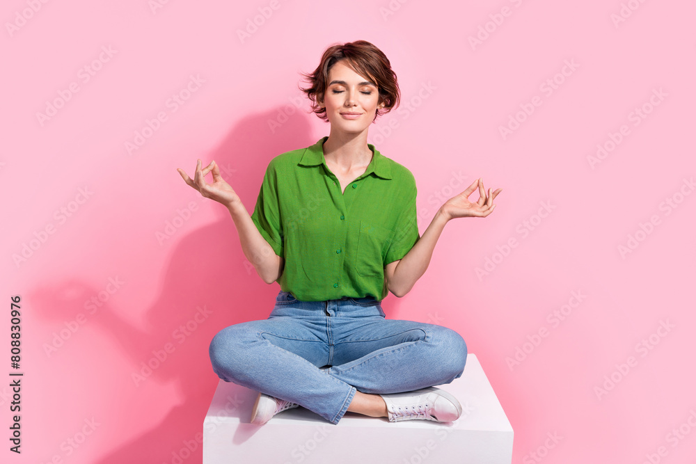 Full size photo of peaceful focused woman dressed green shirt jeans sit on cube in meditation pose isolated on pink color background Full size photo of peaceful focused woman dressed green shirt jeans sit on cube in meditation pose isolated on pink color background