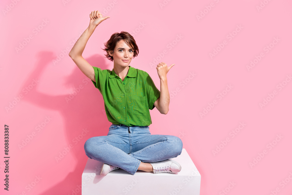 Full size photo of pleasant woman dressed green shirt jeans sit on cube indicating at offer empty space isolated on pink color background Full size photo of pleasant woman dressed green shirt jeans sit on cube indicating at offer empty space isolated on pink color background