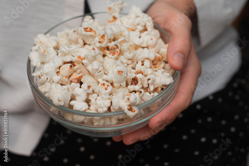 Woman Holding Bowl of Fresh Popcorn. Close-up of a woman's hands holding a transparent bowl filled with freshly popped popcorn, on a natural woven mat.