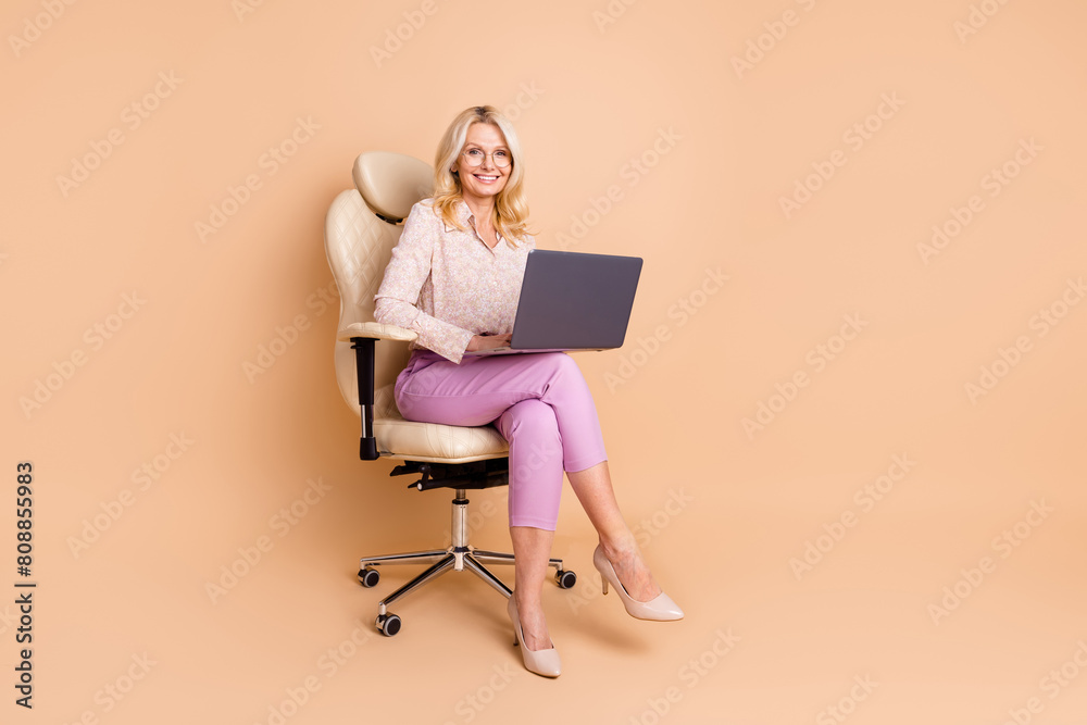 Full body photo of nice retired female sit office armchair hold netbook wear trendy formalwear isolated on beige color background Full body photo of nice retired female sit office armchair hold netbook wear trendy formalwear isolated on beige color background