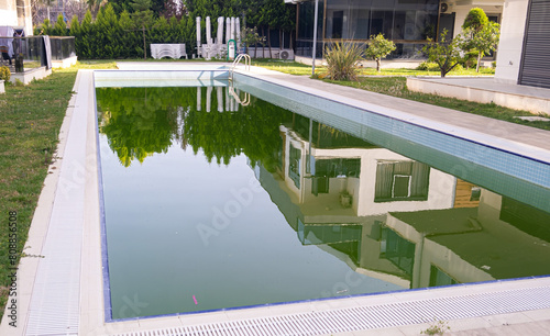 Dirty Swimming Pool with Green Water. A close-up view of a neglected swimming pool with murky green water, reflecting its surroundings.