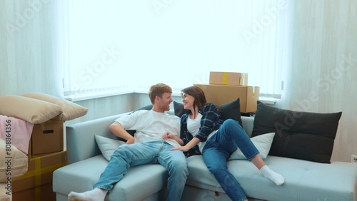 Camera approaching joyful male and pretty female smiling jumping on a sofa in light room with many cardboard boxes. Close up of happy boyfriend and girlfriend moving in together