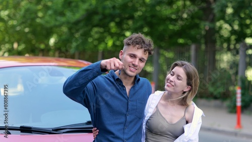 Close up of cheerful young couple man and woman smiling feeling happy with a new car. Caucasian man holding in hand and showing to camera keys to a red automobile. Family purchase.