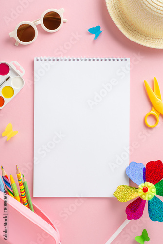 Preschool summer adventure concept. Vertical top view of art supplies - drawing album, colored pencils, paints, hat, glasses, pinwheel, butterflies on light pink surface. Space for advertisement