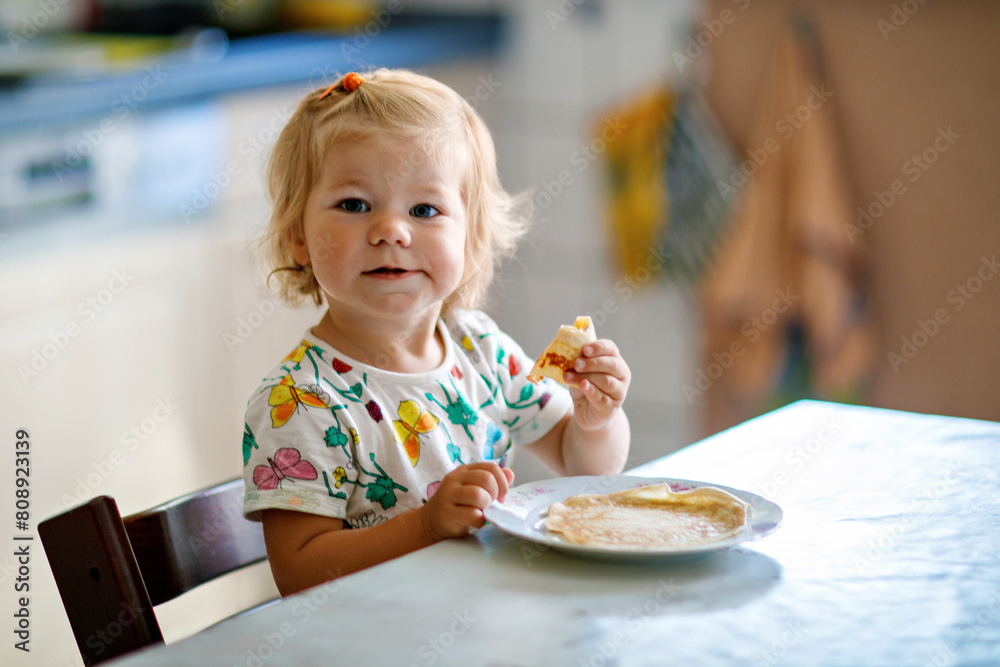 Happy little toddler girl eating delicious pancakes sitting in the kitchen. Cute child tasting different food Happy little toddler girl eating delicious pancakes sitting in the kitchen. Cute child tasting different food