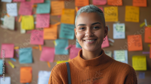 happy non-binary person with short hair looking at camera near corkboard with paper notes in office Stock Photo photography